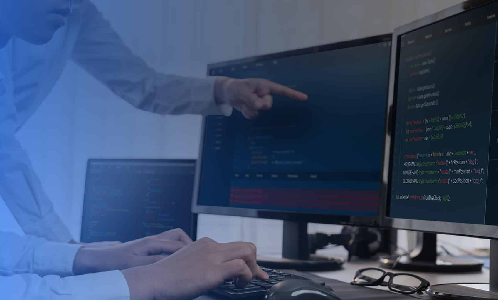Two people work at a desk with multiple monitors displaying code; one types on a keyboard while the other points at the screen, highlighting collaboration and IT Service Desk Solutions. Glasses and a mouse rest on the desk.