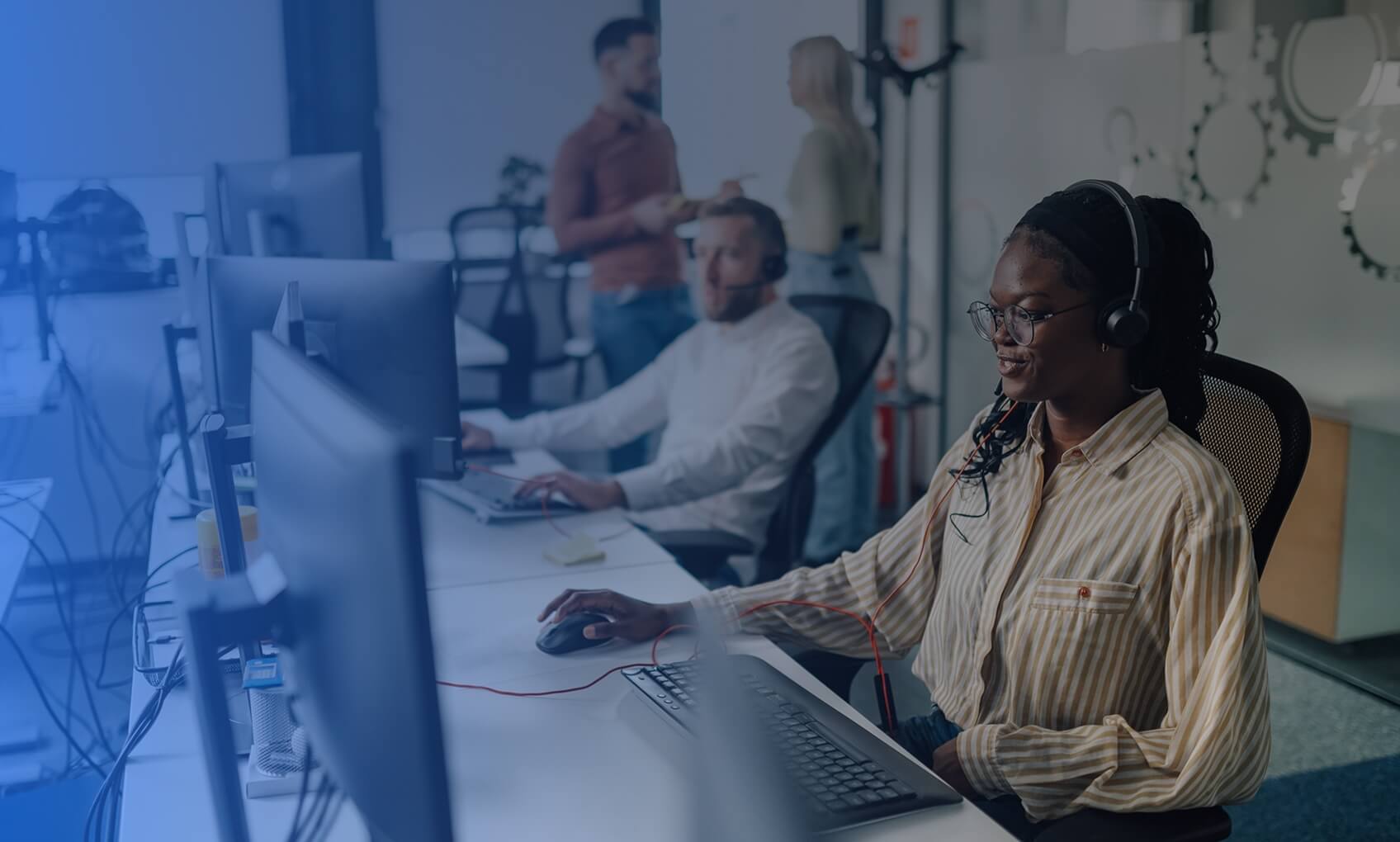 A woman wearing glasses and a headset works at a computer in a modern office. Two colleagues sit nearby, while two others stand and talk in the background. The atmosphere is busy and collaborative.