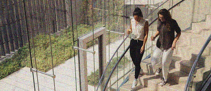Two women holding coffee cups walk up a modern, sunlit staircase with glass railings and a view of greenery outside, discussing innovative IT Service Desk Solutions.