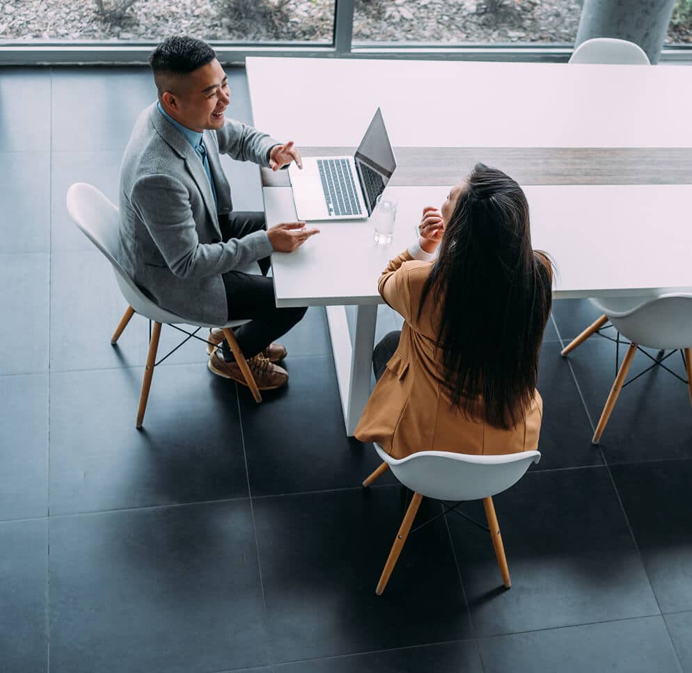 Two people sit across from each other at a modern white table, engaged in conversation. One uses a laptop, while the other listens attentively. The setting is a bright, professional office space with large windows.
