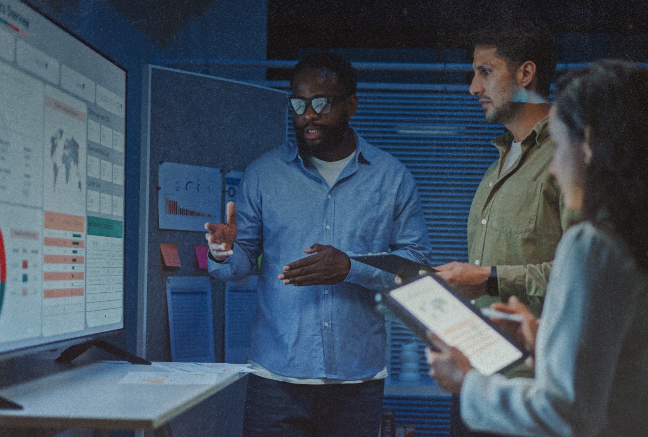Three people stand in a dimly lit office, discussing data displayed on a large screen. One person gestures towards the screen while the others hold a clipboard and tablet, listening and observing the information.