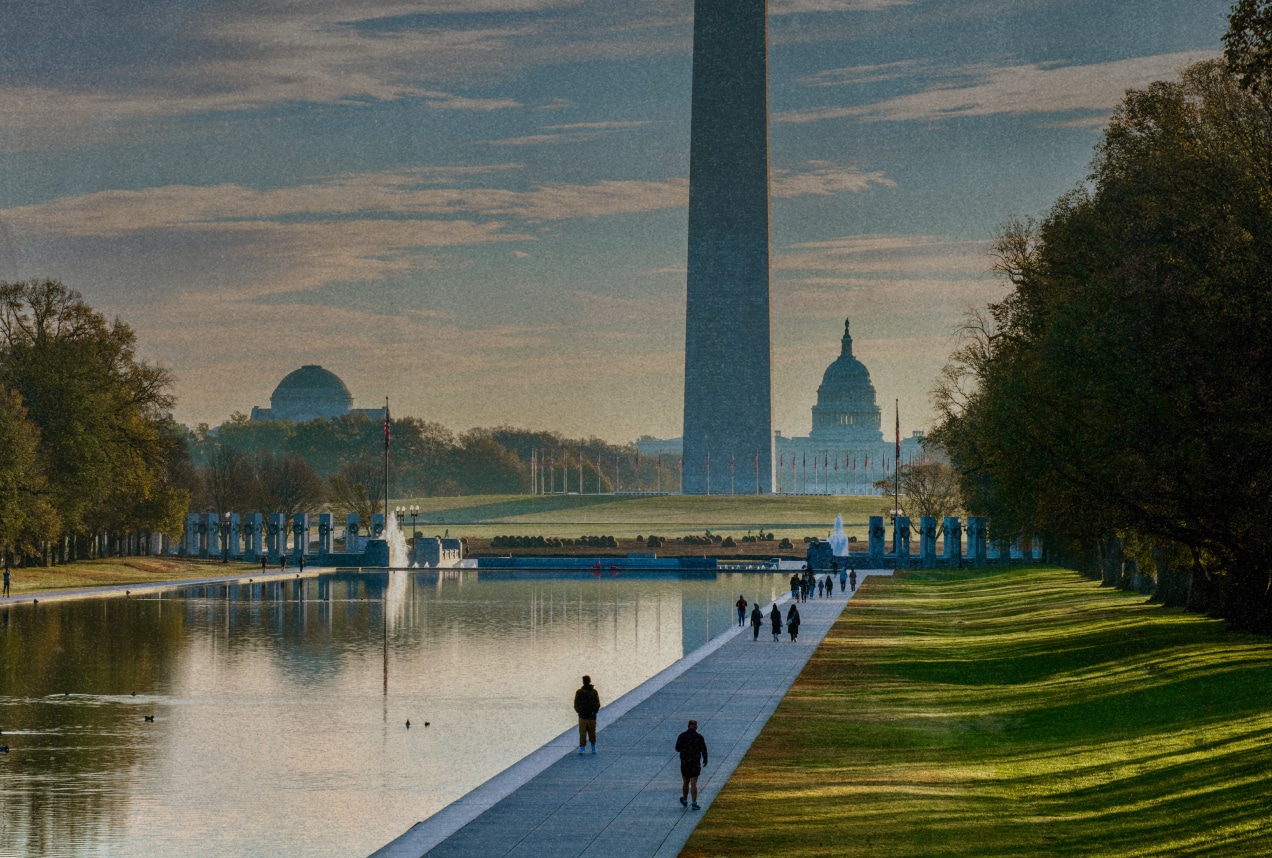 People walk along the Reflecting Pool in Washington, DC, with the Washington Monument in the center and the U.S. Capitol building and other domed structures visible in the background on a partly cloudy day.