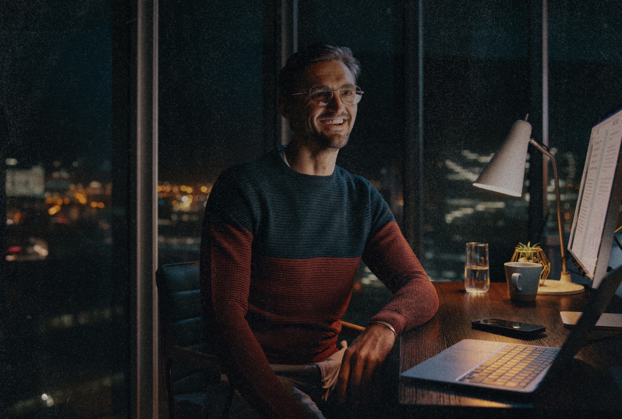 A man wearing glasses and a sweater sits at a desk with a computer, lamp, phone, and glass of water, smiling at night with city lights visible through the window behind him.