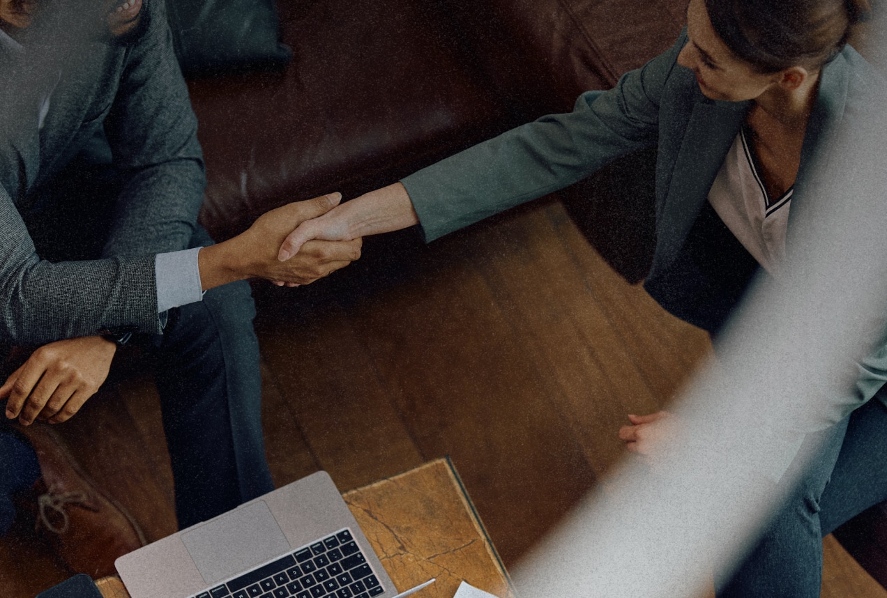 Two people in business attire shake hands across a table with a laptop and documents, suggesting a professional meeting or agreement. The image is taken from above, showing only their upper bodies and hands.
