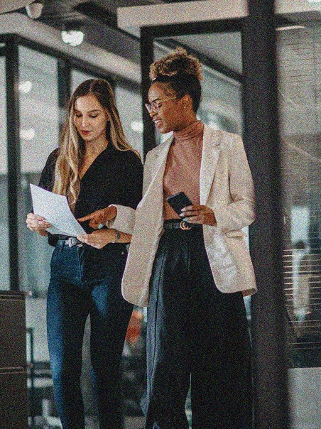 Two women walking in an office hallway, one holding documents and the other holding a smartphone. They appear to be discussing work, both smiling and engaged in conversation.
