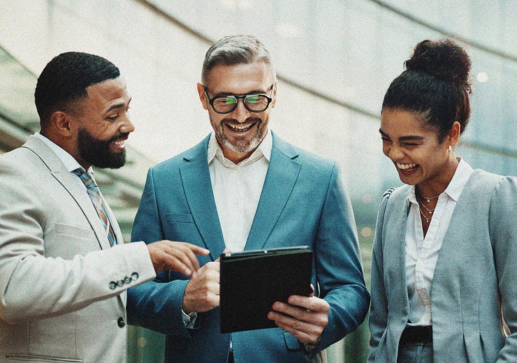 Three business professionals, two men and one woman, stand together indoors, smiling and looking at a tablet one man is holding. They appear to be having a lively, positive discussion in a modern office setting.