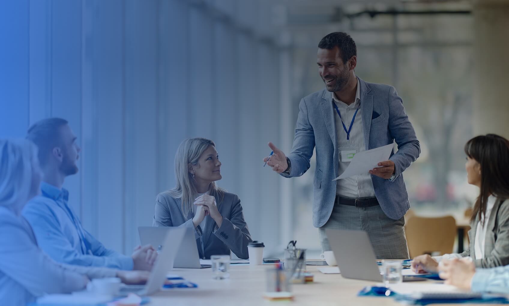 A group of six professionals sit around a conference table discussing a Network Infrastructure Upgrade. One man stands and speaks while holding papers, as others listen attentively. Laptops and documents are on the table, and large windows brighten the room.