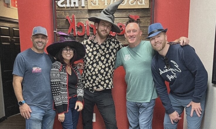 Five smiling adults stand together indoors, three wearing witch hats. The group is casually dressed and poses in front of a red and brown wall with signs in the background.