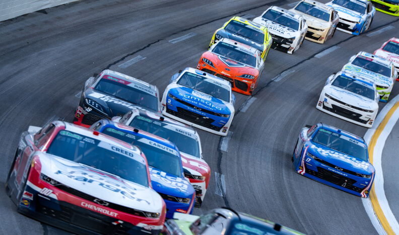 Multiple race cars speed closely together on a paved racetrack, rounding a curve during a NASCAR race. The cars display various sponsor logos and colors, highlighting a fast-paced motorsport competition.