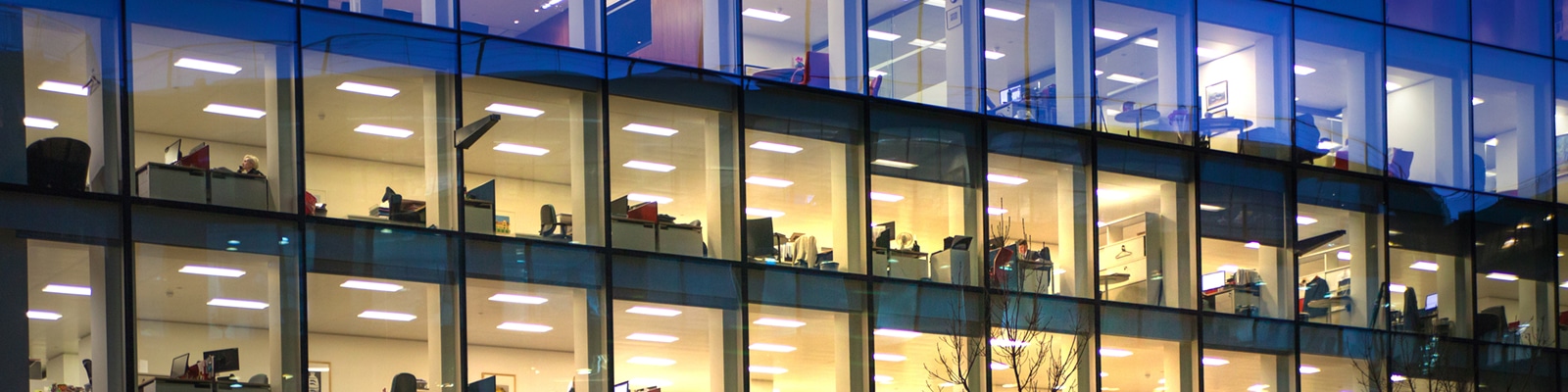 R3 Solutions Governance Risk and Compliance Hero@2x A modern office building at night, with large glass windows revealing rows of brightly lit, empty workspaces—an environment where Governance Risk and Compliance teams work to ensure operational integrity across multiple floors.
