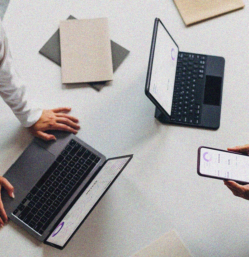 Three people collaborate at a table with two open laptops, folders, and one person holding a smartphone, suggesting a productive work or study environment focused on IT Asset Management.