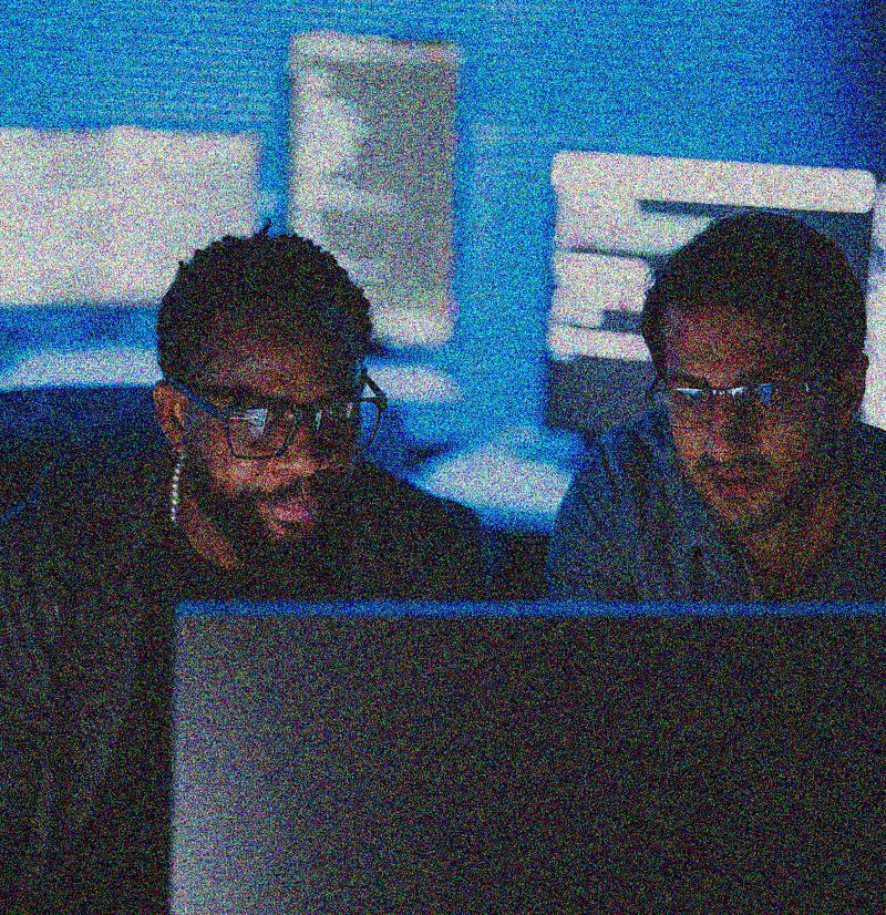 Two men wearing glasses sit side by side in front of a computer screen, illuminated by its light, discussing IT Asset Management amid blurred monitors and blue tones in the background.