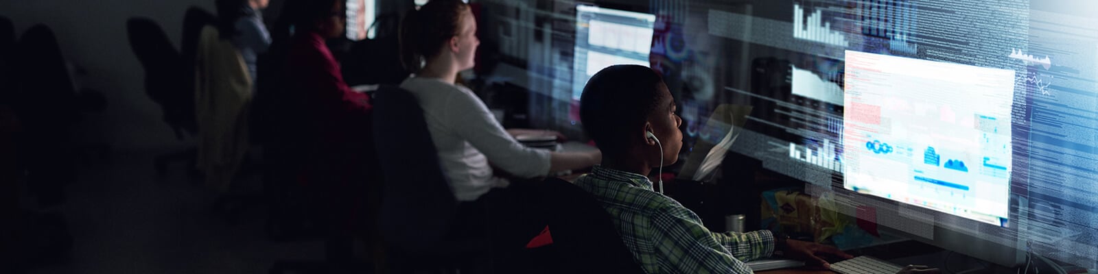Several people sit at desks in a dimly lit room, working on desktop computers with data and IT Asset Management dashboards displayed on the monitors. The environment looks like a modern office or technology lab.