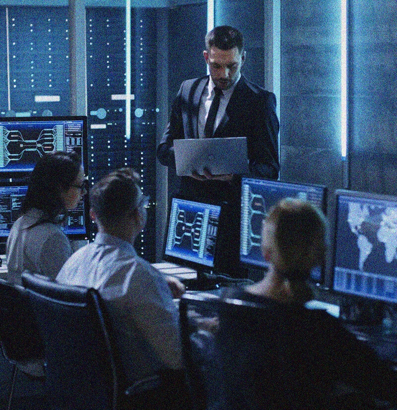 Four people in a dark, modern control room with multiple computer screens display data and maps related to IT Asset Management. One person stands holding a laptop, while three others sit facing the monitors, appearing focused on their work.