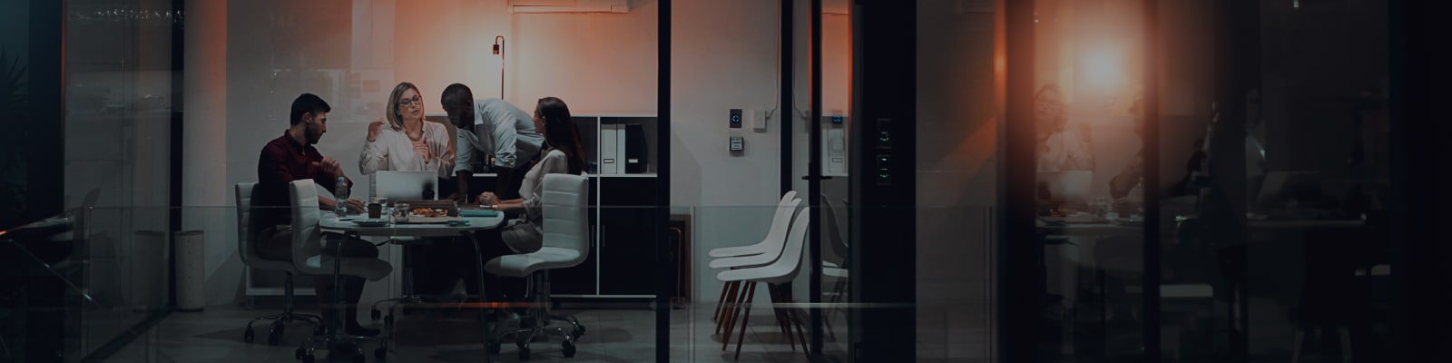 A group of five people sit and stand around a conference table in a modern office, engaged in discussion, with laptops and documents on the table and glass walls surrounding the room.