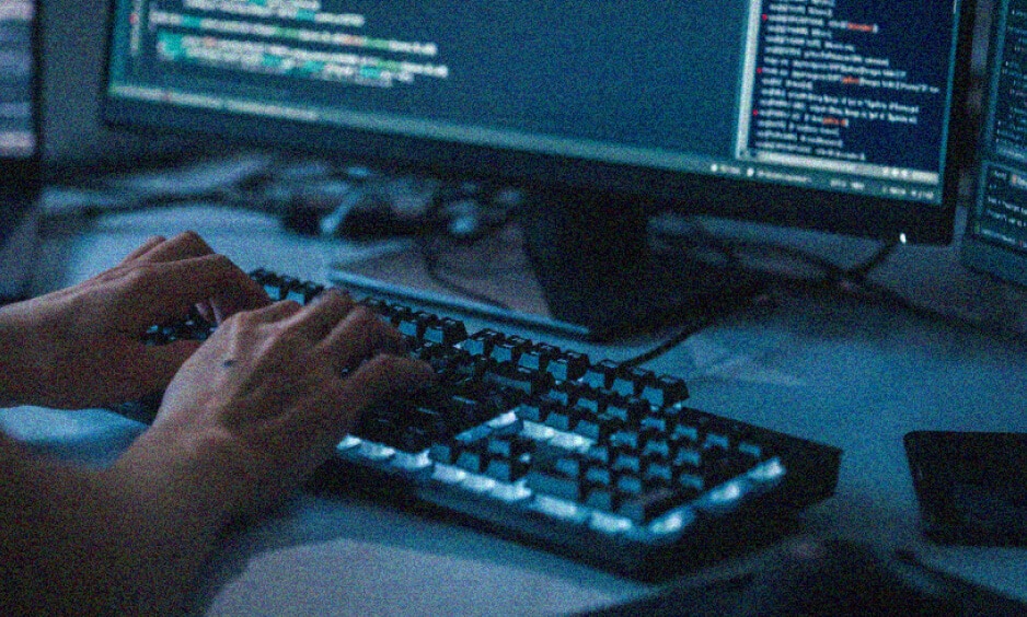 Hands typing on a mechanical keyboard in front of a computer monitor displaying lines of code, suggesting programming or software development work in a dimly lit environment focused on delivering IT solutions.