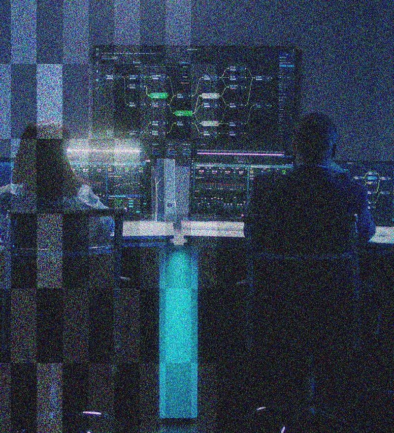 Two people sit at computer desks in a dark room, facing a large, illuminated screen displaying complex digital data and network diagrams—capturing the high-tech atmosphere of an IT Solutions control center.