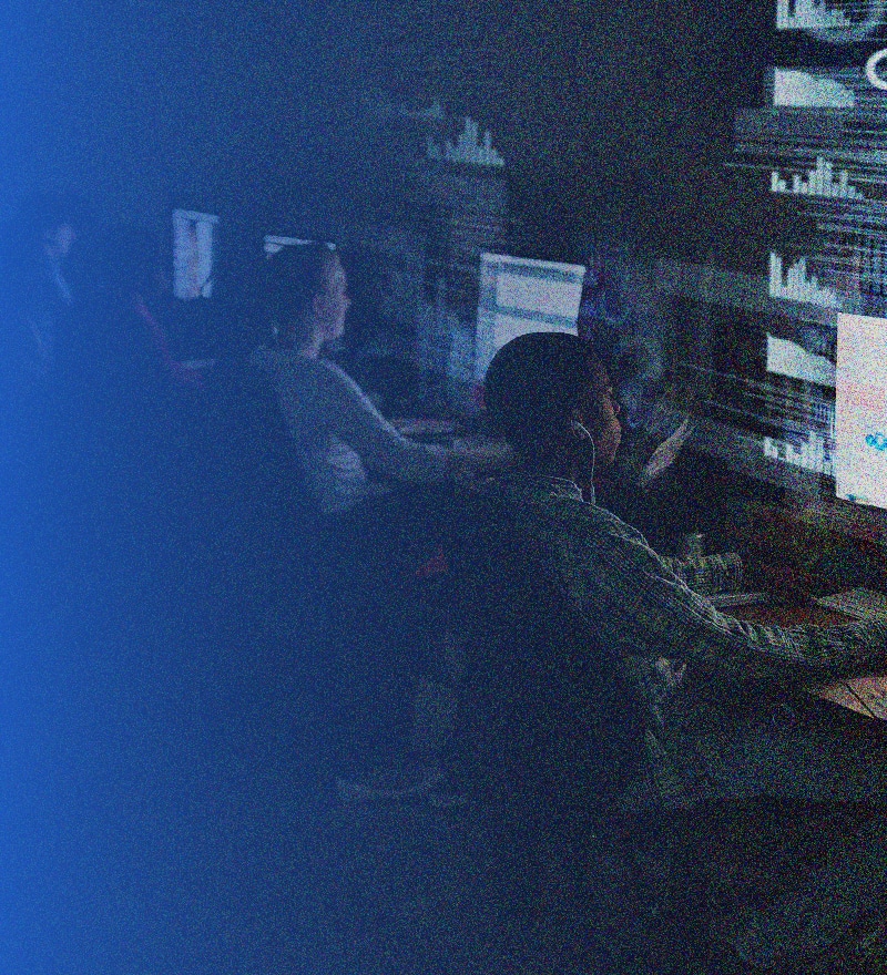 Several people sit at computer desks in a dark room, illuminated by the glow of monitors displaying graphs and data visualizations—a focused, high-tech environment dedicated to advanced IT solutions.