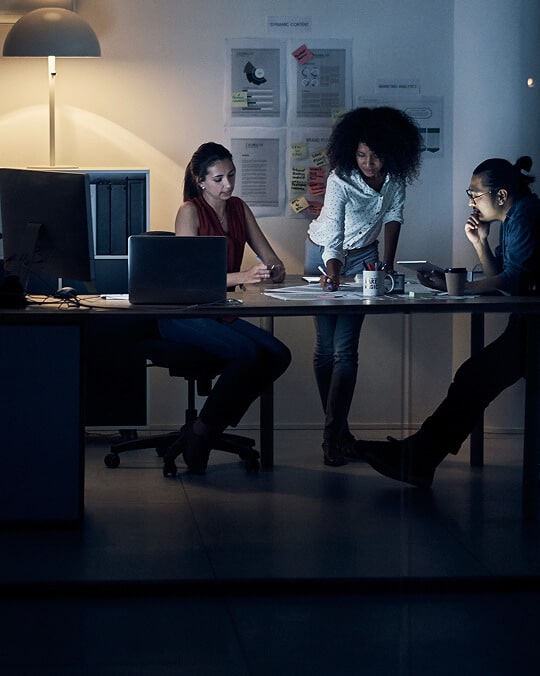 Three people work together in a dimly lit office at night, gathered around a desk with laptops and documents, while notes and charts are pinned to the wall behind them.