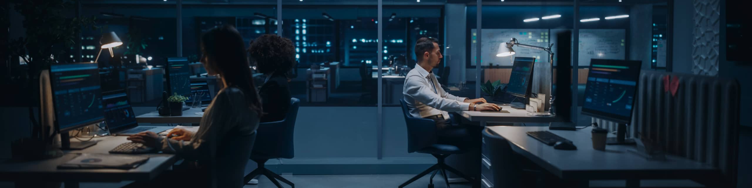 Several people work late at desks in a modern office, illuminated by desk lamps and computer screens, with large windows showing city lights outside.