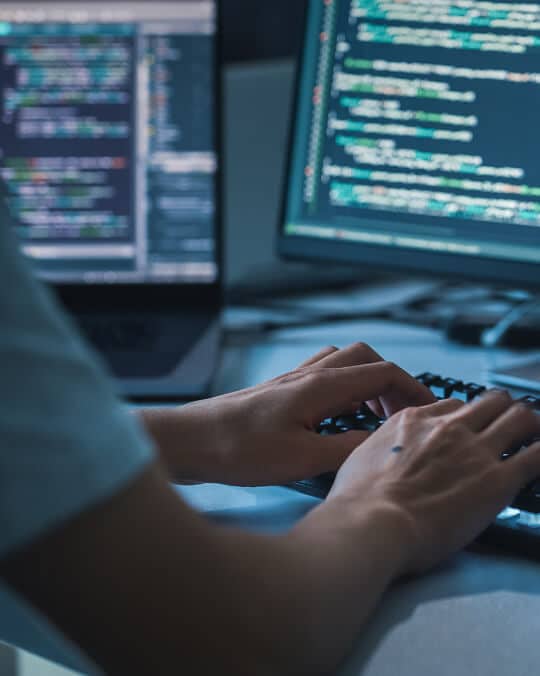 A person types on a keyboard in front of two monitors displaying lines of code, suggesting programming or software development work in a dimly lit environment.