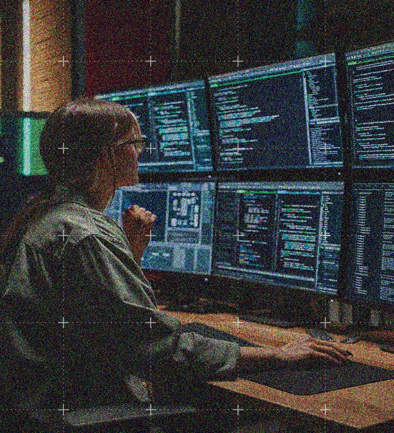 A person with long hair tied back sits at a desk, looking at multiple large monitors displaying lines of code and data in a dimly lit room, suggesting a programming or IT Solutions environment.