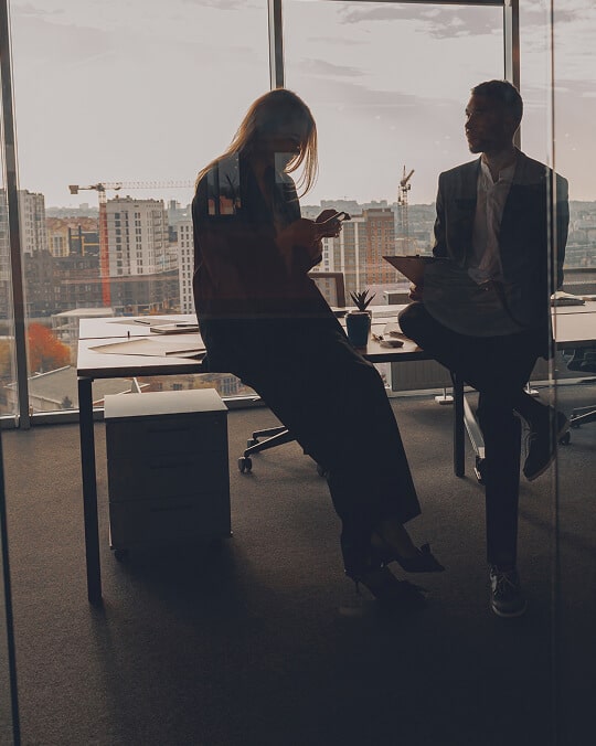 Two people in business attire sit and stand by a desk in a modern office with large windows overlooking a cityscape. One is using a phone, and the other holds a tablet. The atmosphere is dimly lit.