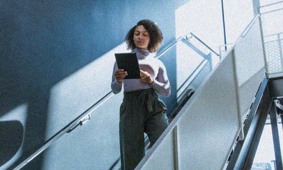 A woman in a turtleneck and high-waisted pants walks down a staircase, engrossed in her tablet—perhaps reviewing the latest IT solutions as bright sunlight casts striking shadows on the wall behind her.