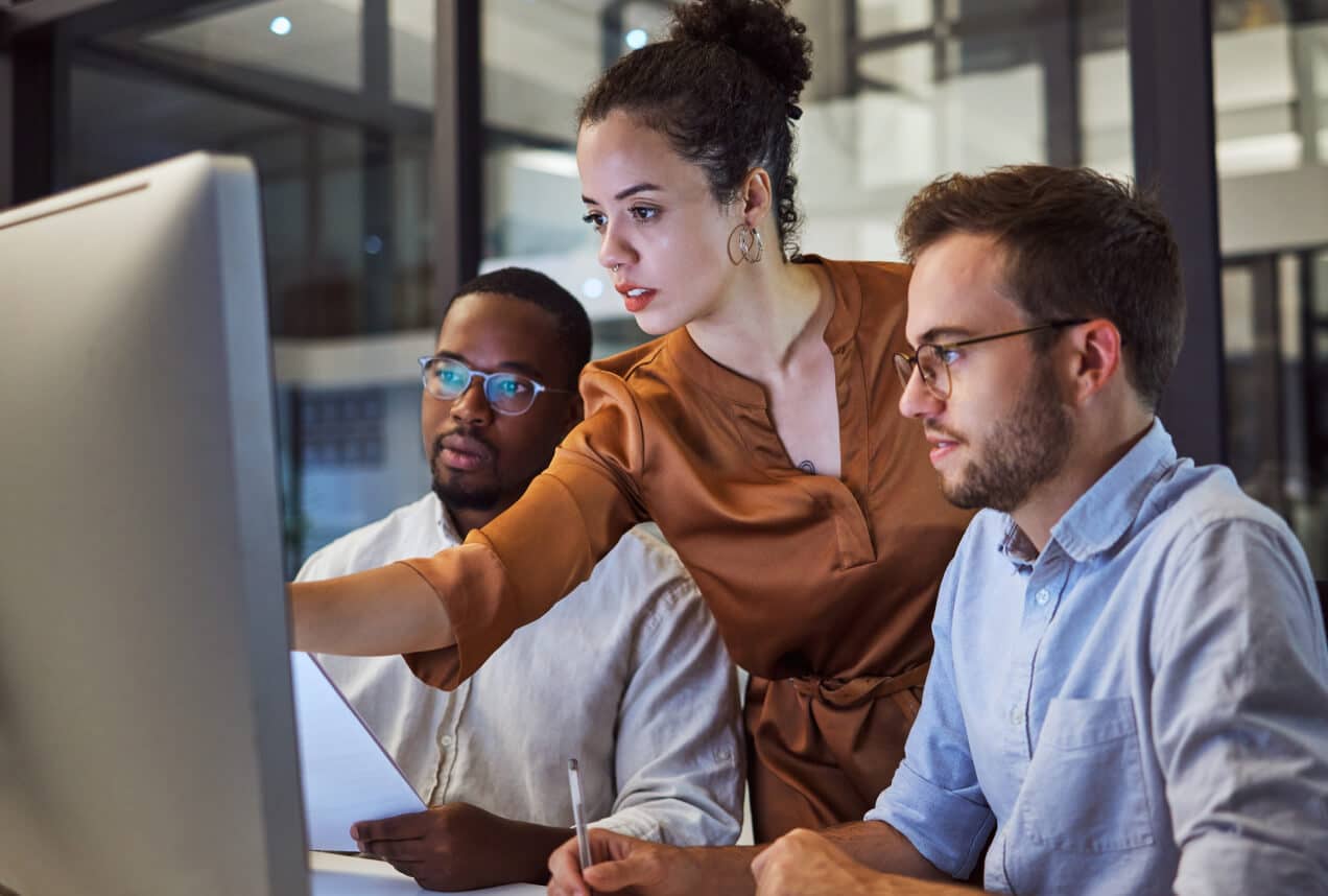 Three people work together at a computer in an office. A woman stands, pointing at the screen, while two men sit, focused on the monitor. They appear to be collaborating on a project.