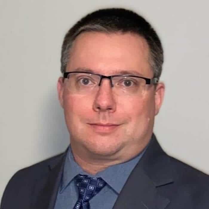 A man with short dark hair and glasses is wearing a dark suit jacket, a blue dress shirt, and a patterned blue tie, posing in front of a plain light-colored background.