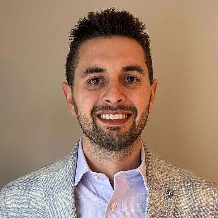 A man with short brown hair and a beard, wearing a light gray plaid blazer and a light purple shirt, smiles in front of a plain beige background.