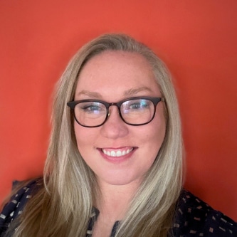 A woman with long blonde hair and glasses smiles at the camera. She is wearing a dark, patterned top and is standing in front of a bright orange background.