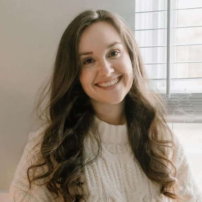 A woman with long brown hair, wearing a white knit sweater, smiles while sitting indoors near a window with blinds. Soft natural light brightens the room.