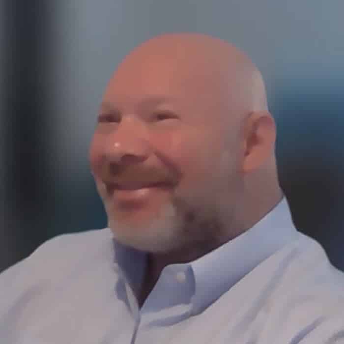 A bald man with a trimmed beard, wearing a light blue collared shirt, smiling and looking to the side against a blurred background.