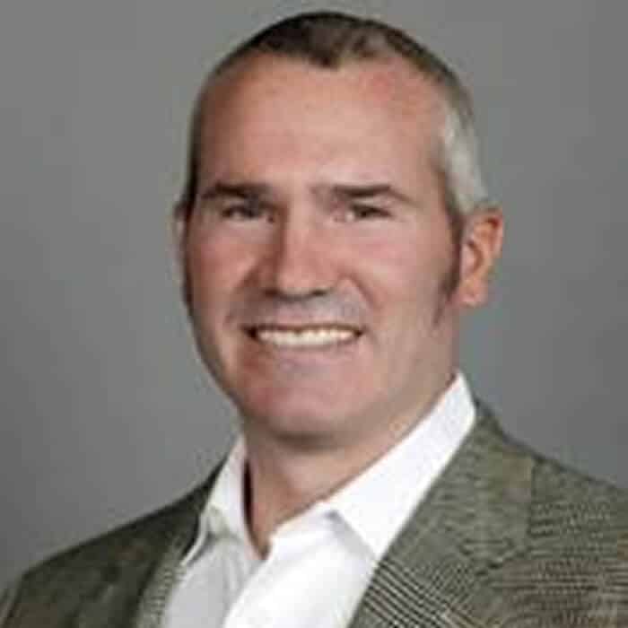 Smiling man with short gray hair wearing a light-colored dress shirt and a gray textured blazer, posing in front of a plain gray background.