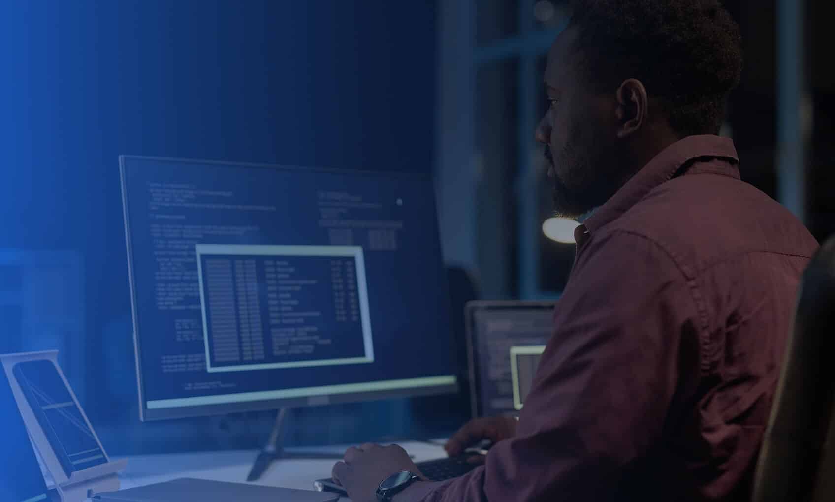 A person sits at a desk using multiple monitors displaying code and command line interfaces in a dimly lit room.