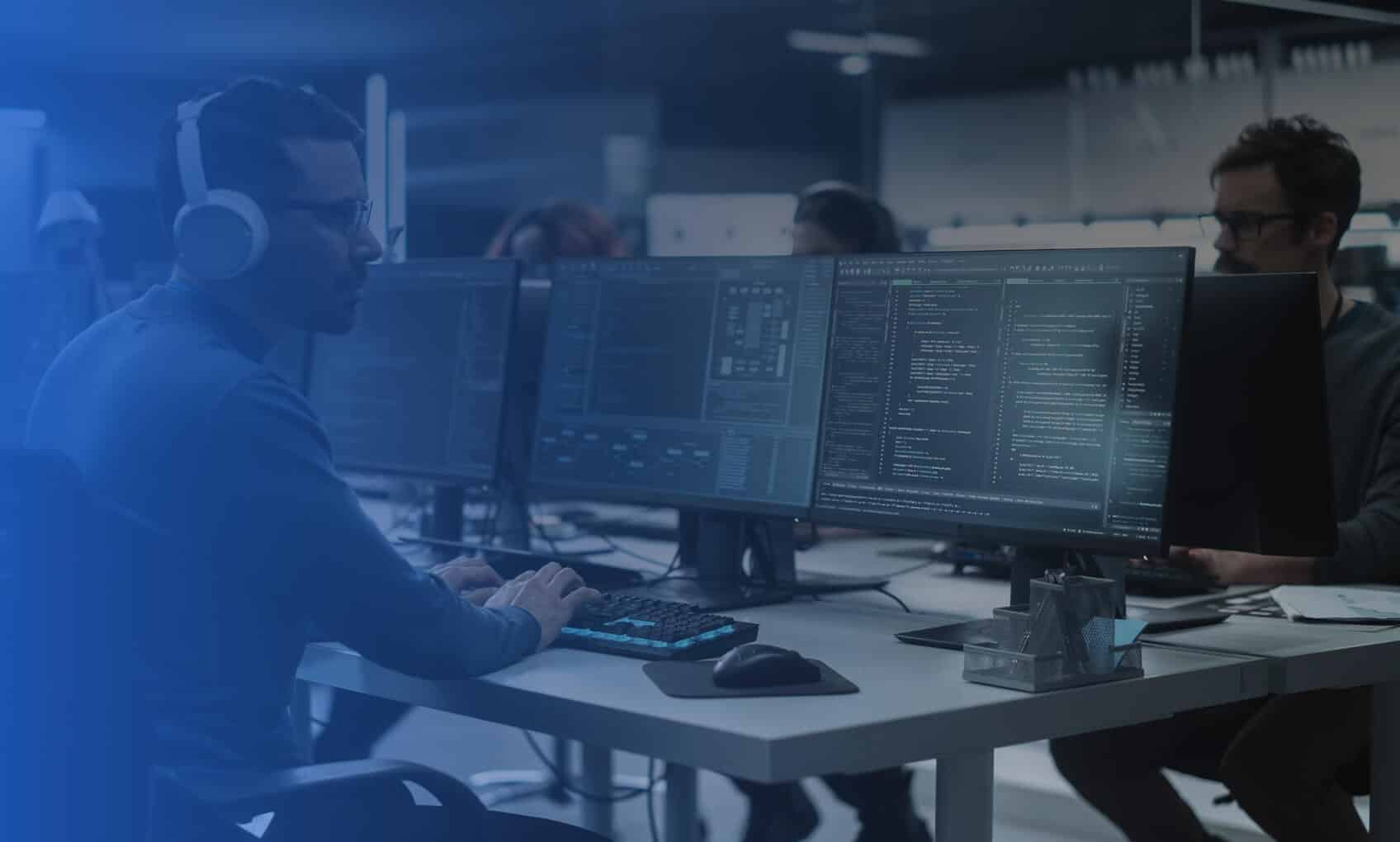 Several people work at desks with multiple large monitors displaying code in a modern, dimly lit office; one person wears headphones and types on a keyboard.