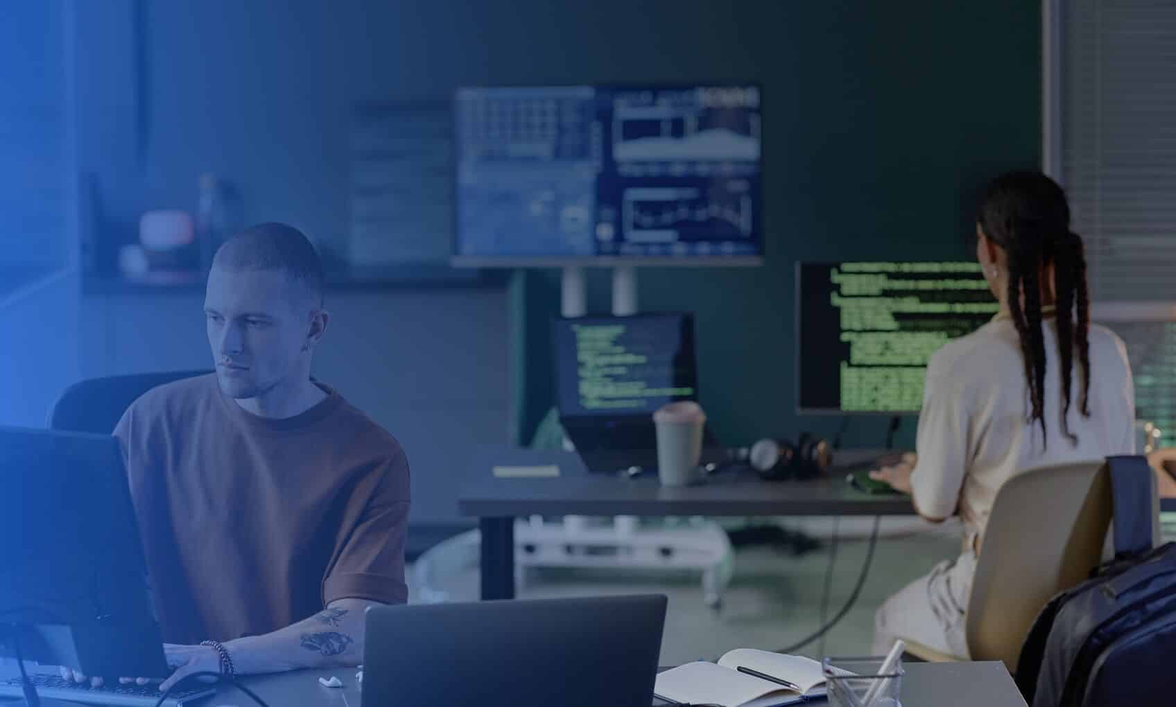 Two people work at computer desks in an office. One person faces the camera, while the other reviews green text related to Azure Sentinel Implementation on a monitor. Multiple screens and office supplies are visible in the background.