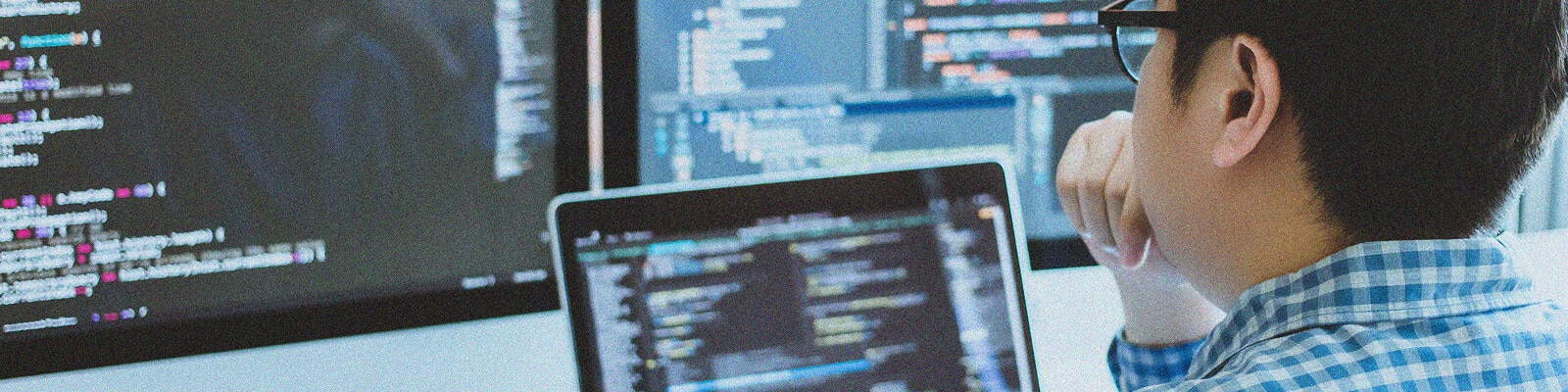 A person wearing glasses and a checkered shirt is seated at a desk, focused on multiple computer screens displaying lines of code while working on a Teams Calling Implementation.