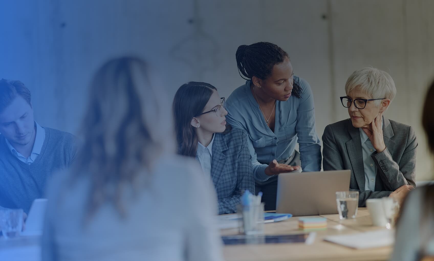 A diverse group of professionals discusses Azure Sentinel Implementation around a conference table; one woman stands and gestures as others listen, take notes, and use laptops.