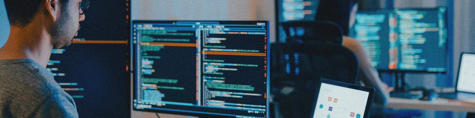 R3 Technology Assessment@2x Two people sit at desks working on computers with multiple monitors displaying lines of code, programming interfaces, and Technology Assessment reports in a dimly lit office or workspace.