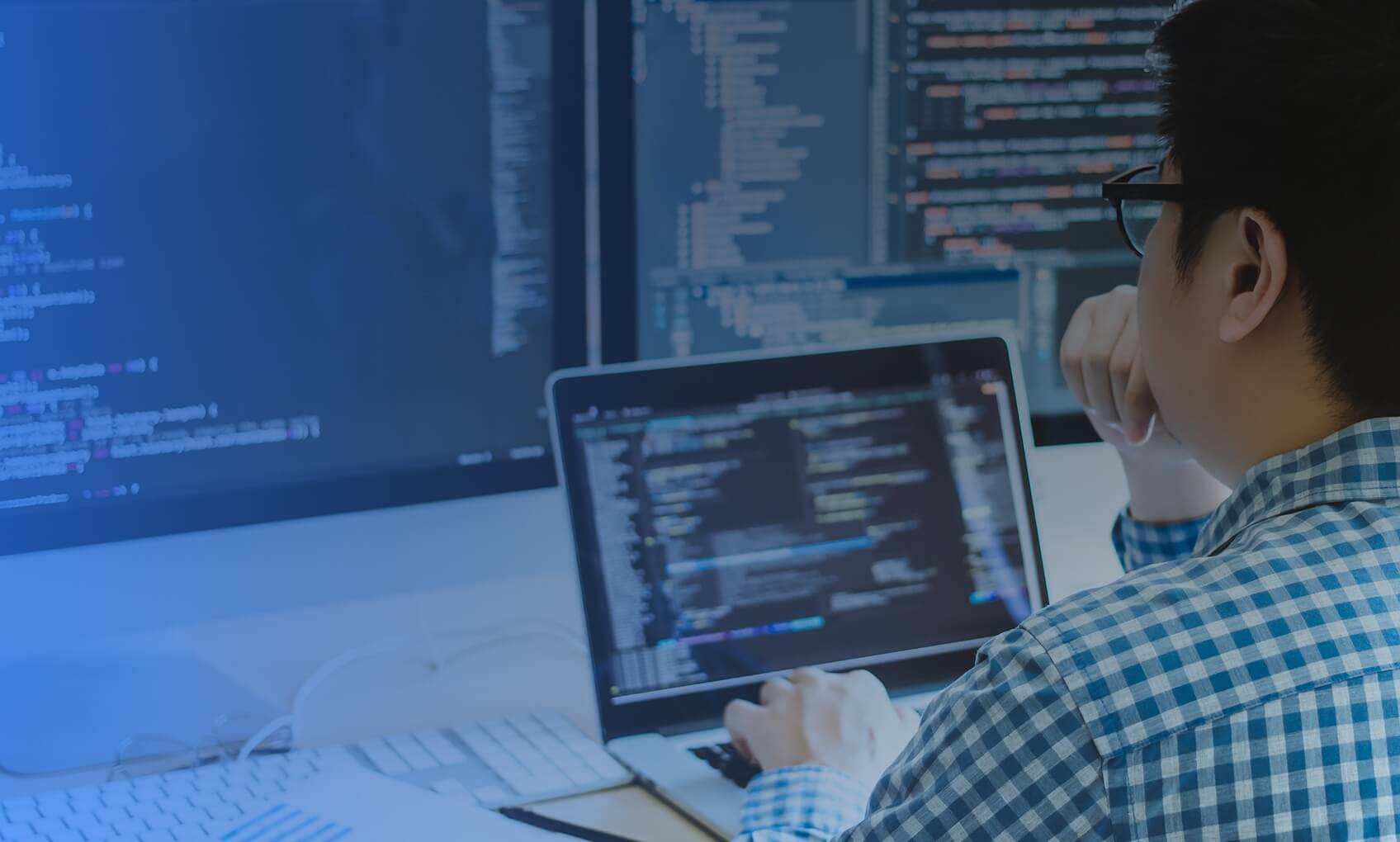 A person in a blue checkered shirt sits at a desk, working on a laptop with code on the screen. Two large monitors displaying programming code related to Azure Sentinel Implementation are in front of them.