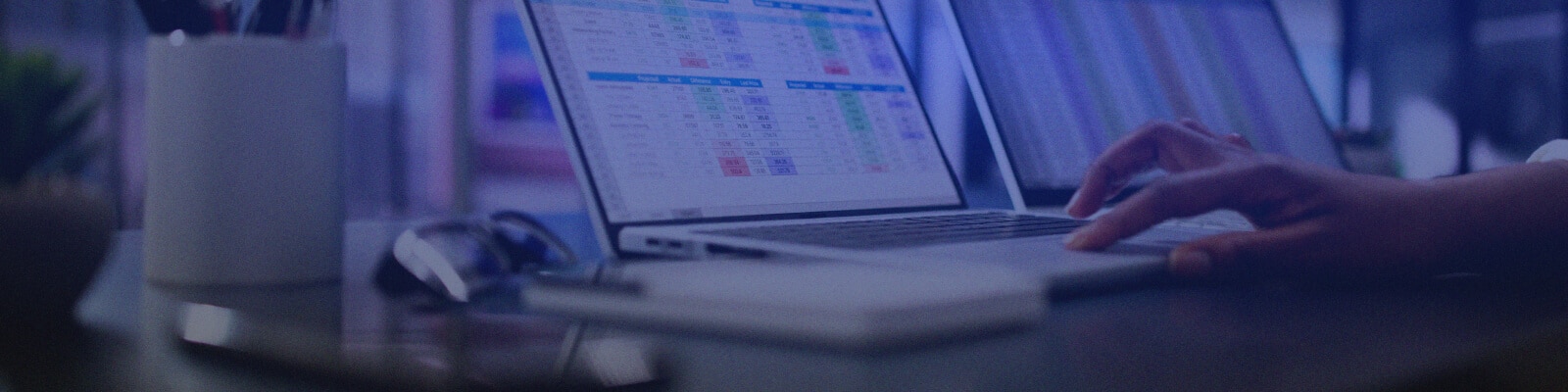 A person uses a laptop displaying colorful spreadsheets at a dimly lit desk, planning Wireless Infrastructure Implementation, with a notepad, pen holder, and glasses nearby.