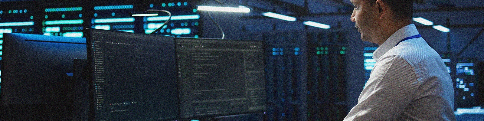 A person in a white shirt stands in a server room, observing two computer monitors displaying code and Zero-Trust Architecture details, with blue-lit server racks glowing in the background.