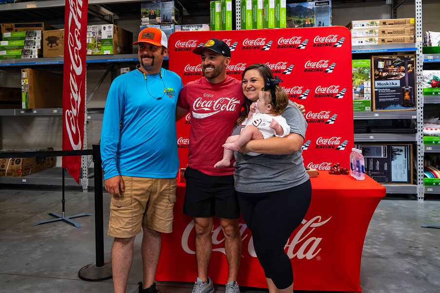Four people, including a baby, pose and smile in front of a red Coca-Cola backdrop and table inside a store with shelves stocked with boxes and products.