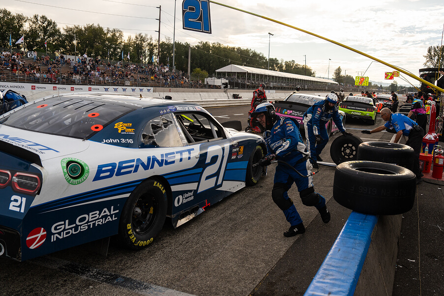 A race car marked “21” is in a pit stop area as crew members in blue uniforms change its tires. Other pit crews work on nearby cars, while spectators watch from the stands in the background.