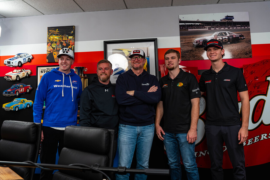 Five men stand together in a room decorated with racing memorabilia, model cars, and racing photos on the wall behind them. They are smiling and wearing casual clothing, including branded shirts and hats.