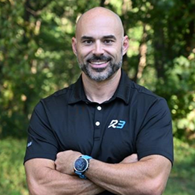 A smiling bald man with a beard stands outdoors in front of trees, wearing a black polo shirt with R3 on it and a smartwatch on his wrist, arms crossed confidently.