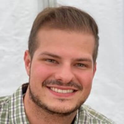 Smiling man with short brown hair and a trimmed beard, wearing a green and white plaid shirt, in front of a white background.